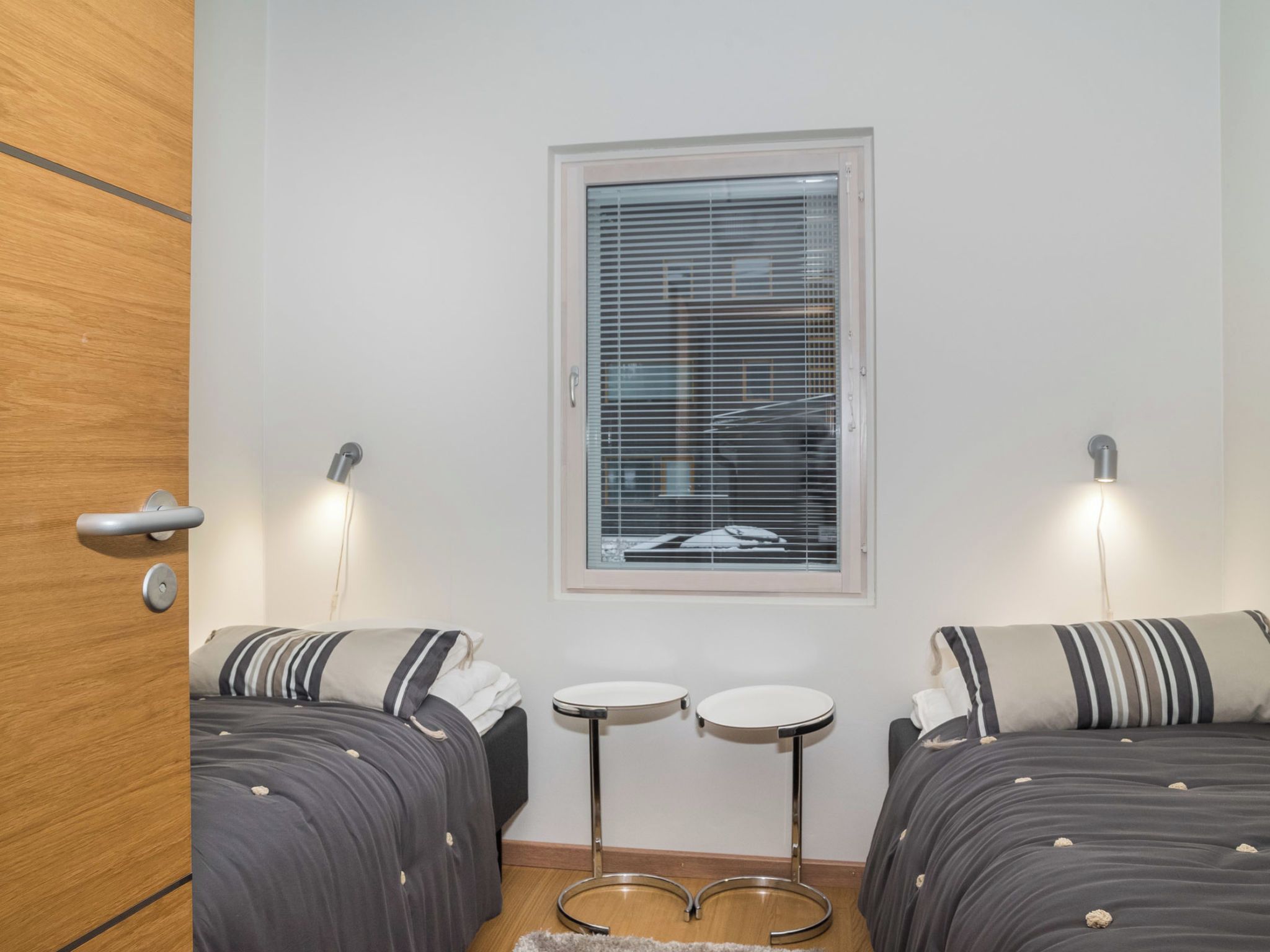 Una habitación con dos camas, un ventanal con persianas, dos mesitas pequeñas y una puerta.