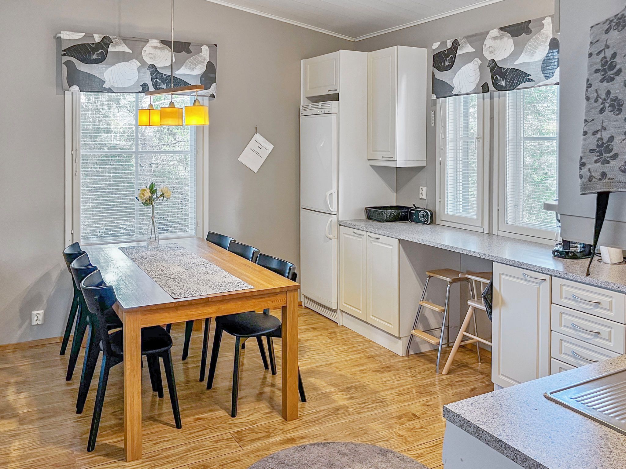Una cocina moderna con armarios blancos, una mesa de comedor de madera con sillas y una ventana con cortinas decorativas.