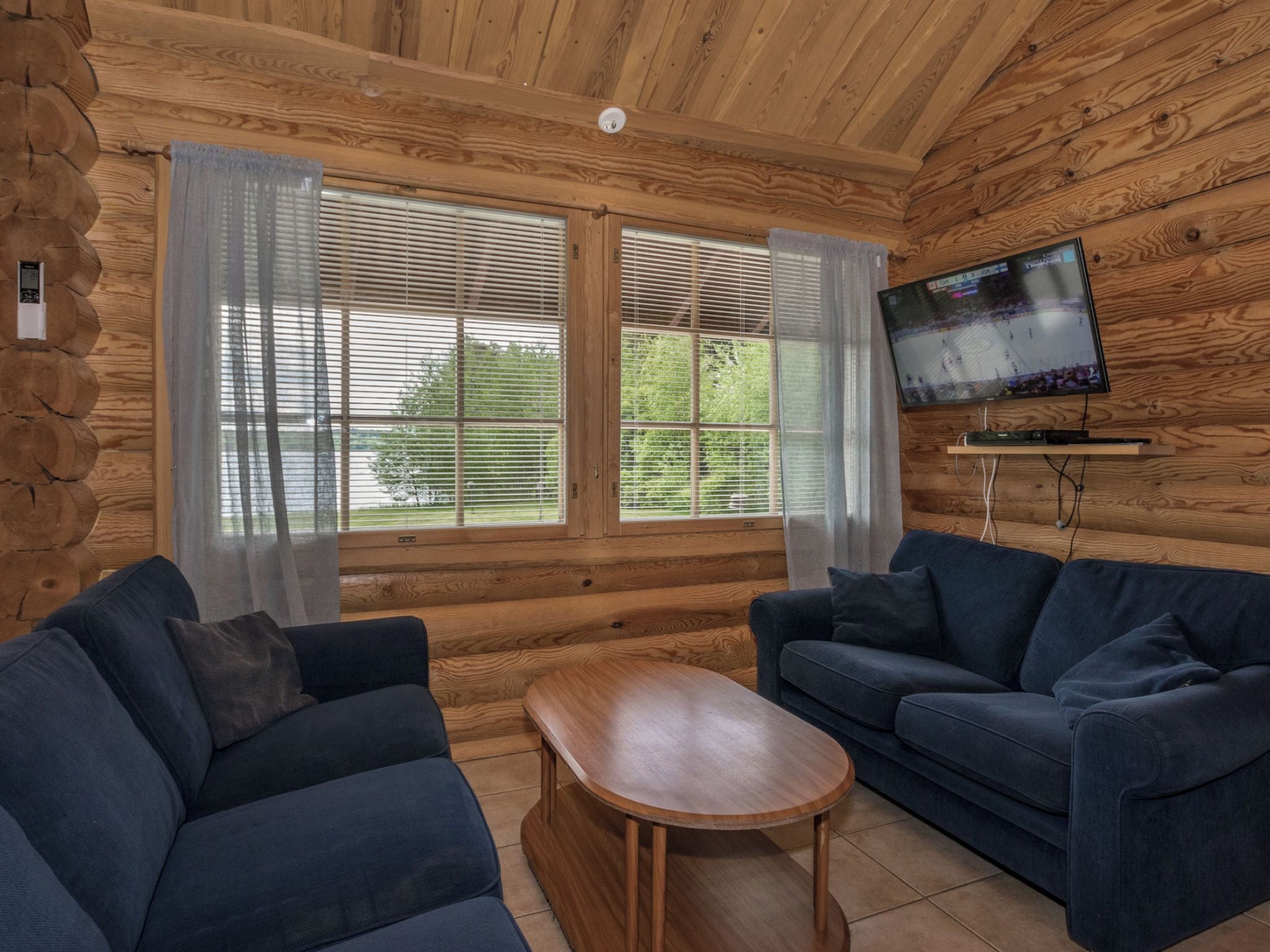 A log cabin interior with two sofas, a coffee table, a TV mounted on the wall, and large windows with curtains.