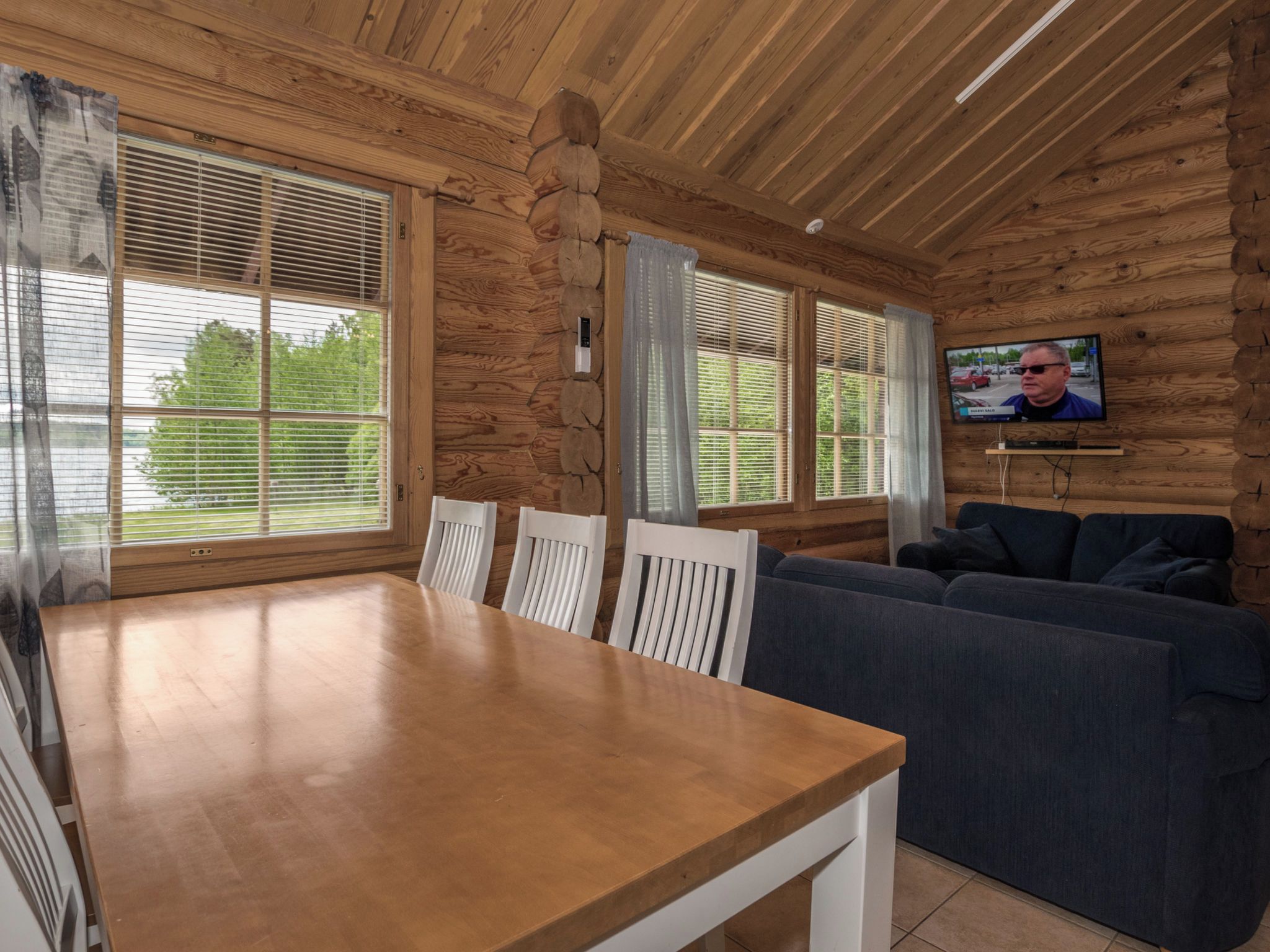 A wooden interior with a dining table, white chairs, a dark sofa, and a television mounted on the wall.