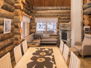 A log cabin interior with a stone fireplace, a sofa, a dining table, and a window with a view of trees.