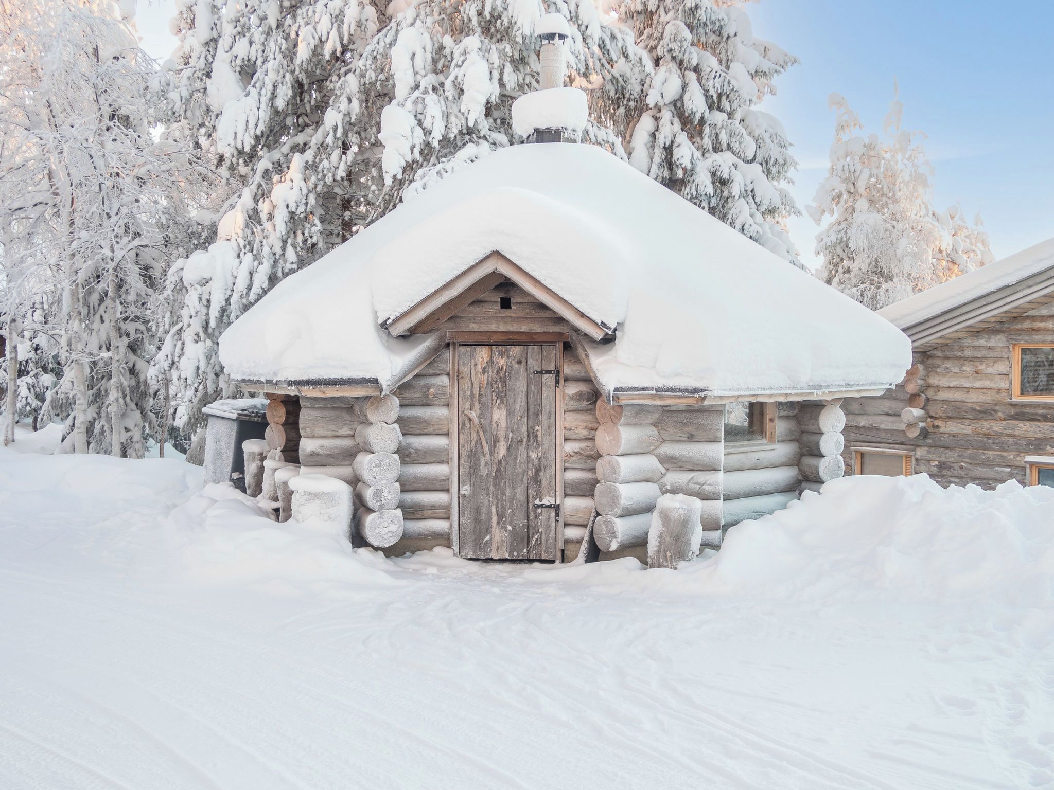 Eine Blockhütte mit schneebedecktem Dach und einer Holztür, umgeben von Schnee und Bäumen.