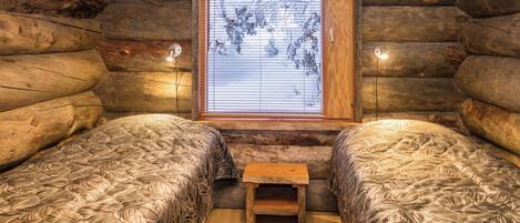 A log cabin room with two beds, a small wooden bench, and a window with blinds.