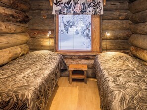 A log cabin room with two beds, a small wooden bench, and a window with blinds.