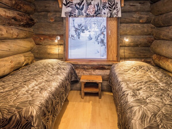 A log cabin room with two beds, a small wooden bench, and a window with blinds.
