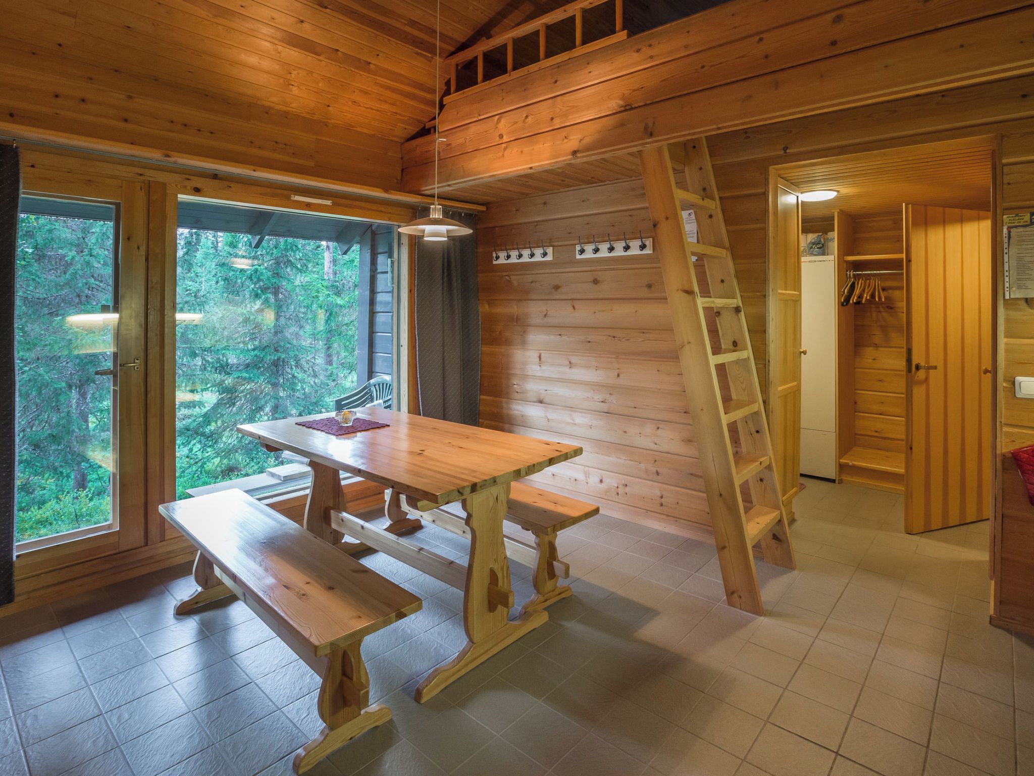 A wooden cabin interior with a dining table and benches, a ladder, and a view of the forest through large windows.
