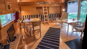 Een gezellig cabin interieur met houten plafond, een eethoek met tafel en stoelen, een bank met gestreepte kussens en een groot raam met uitzicht op de natuur.