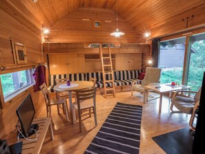 Ein gemütliches Blockhaus-Interieur mit Holzvertäfelung, einem runden Esstisch, einer Couch, einer Hockerleiter und Blick ins Freie.