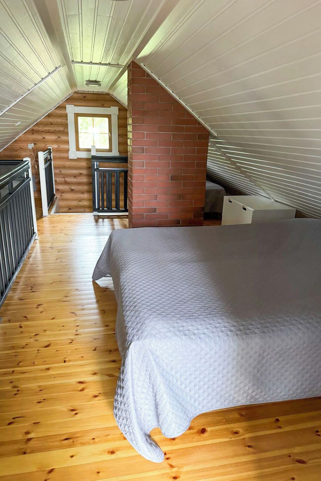 Een slaapkamer met een schuine kap, houten vloer, een bed, een raam en een open haard van baksteen.