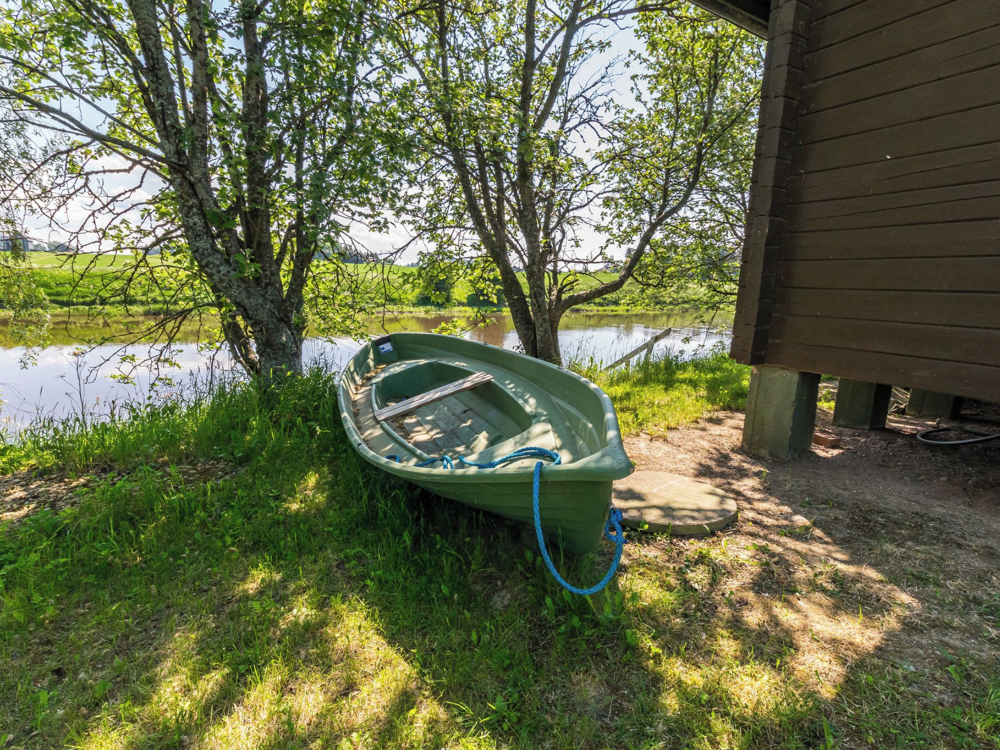 A green boat tied to a tree by a body of water, with a wooden structure in the background.