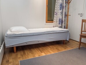 Una cama individual con colchón, almohadas y una manta en una habitación con suelo de madera, una puerta y una ventana con persianas.