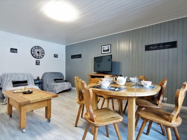 A cozy living room with a dining table, chairs, a sofa, a TV, and a clock on the wall.