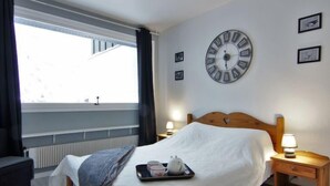 Hotellihuone, jossa on sänky, suuri seinäkello, ikkuna, jossa on verhot, sekä tarjotinalusta, jossa on kuppeja ja teekannu sängyn päällä.