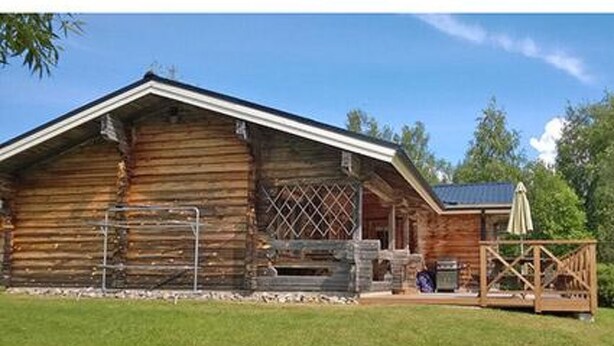Un chalet rustique en rondins de bois, doté d’un porche et d’un espace extérieur aménagé pour s’asseoir.