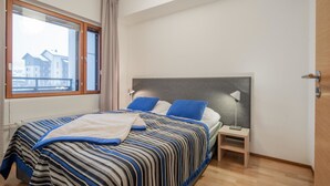 Une chambre à coucher avec un lit, une table de chevet, une lampe, une fenêtre avec des rideaux et une vue sur les bâtiments à l’extérieur.