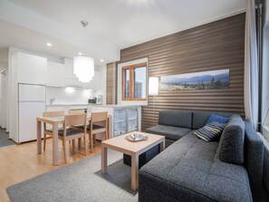 Eine moderne Küche mit weißen Schränken, ein Essbereich mit Tisch und Stühlen, ein graues Sofa mit Blick auf eine Berglandschaft und eine Holzvertäfelung.