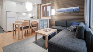 Un salon moderne avec un canapé gris, une table à manger en bois et des chaises. On aperçoit une cuisine avec des meubles blancs et une fenêtre à l’arrière-plan.