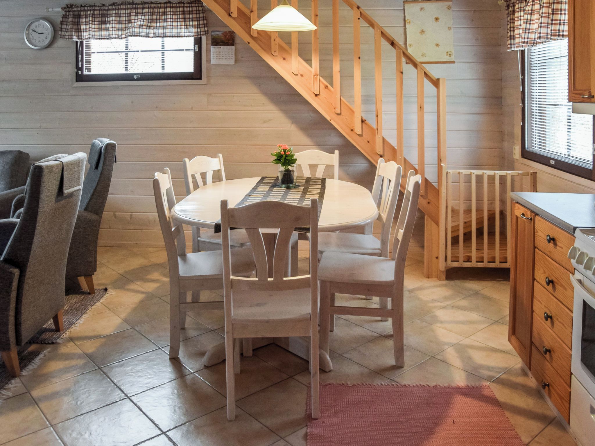 A cozy kitchen with a wooden staircase, a dining table with chairs, a clock, and a window with blinds.
