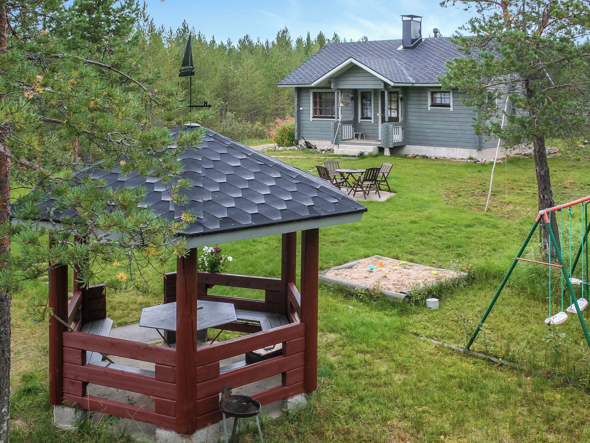 En lusthus med ett sluttande tak, ett utebord och stolar på en gräsplätt, med ett hus i bakgrunden.
