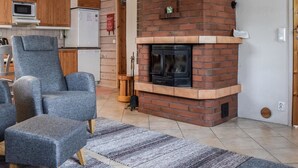 Una sala de estar con chimenea de ladrillo, un sillón gris, un pequeño otoman y una zona de cocina con gabinetes de madera y un refrigerador blanco.