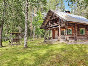 A rustic log cabin with a porch and a small adjacent structure, surrounded by a forest.
