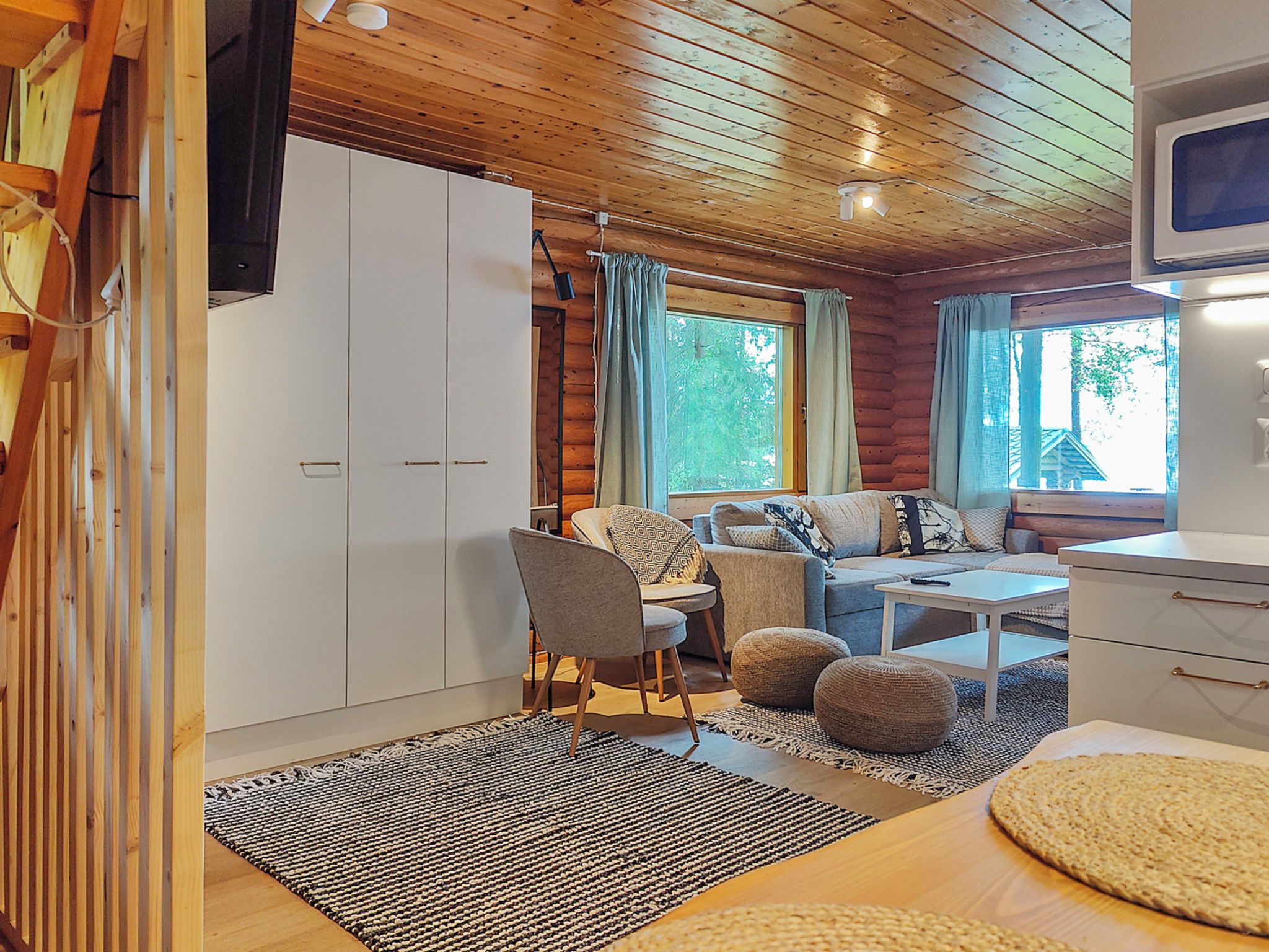 A cozy cabin interior with a wooden ceiling, a sofa set, a dining table, and a TV mounted on the wall.