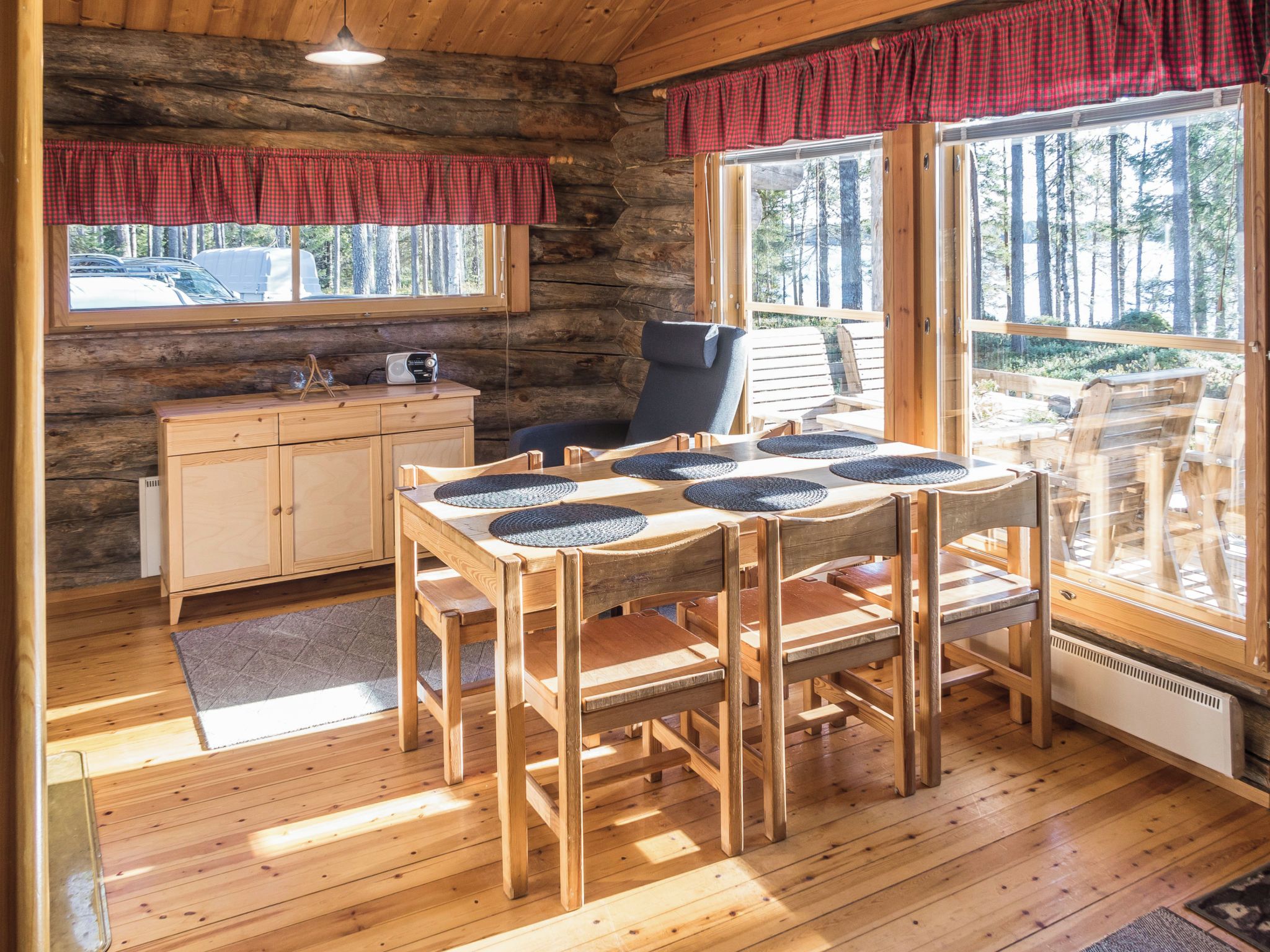 Interior de una cabaña con una mesa de comedor de madera, sillas, un radio y vistas del bosque a través de ventanas amplias.