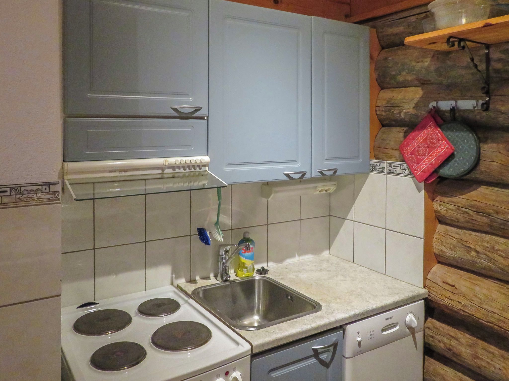 Cocina compacta con estufa, pileta y lavavajillas, con alacenas azul claro y un fondo de pared de piedra.