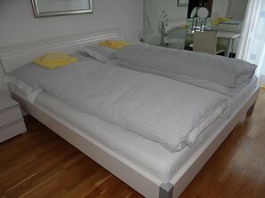 Een tweepersoonsbed met een wit frame en grijze beddengoed, een nachtkastje en een spiegel aan de muur.