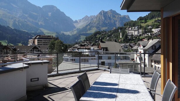 A terrace with a table and chairs, overlooking a mountainous landscape with buildings in the background.