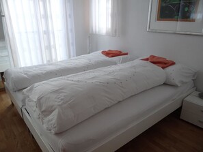 A double bed with white bedding and orange pillows in a room with wooden flooring and white walls.