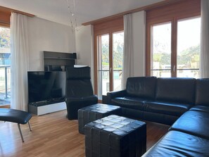 A modern living room with a flat-screen TV, a black leather sofa, a black leather chair, and a wooden floor.