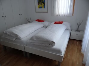 Ein Doppelbett mit weißer Bettwäsche und roten Kissen, ein Nachttisch, ein Bild in einem Rahmen an der Wand und ein Fenster mit durchscheinenden Vorhängen.