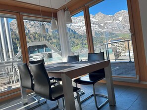 Een eetgedeelte met een tafel en stoelen, grote ramen met uitzicht op bergen, en hangende hanglampen.