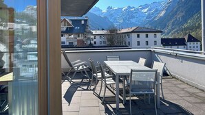 A terrace with outdoor seating, a view of snow-capped mountains, and traditional buildings in the background.
