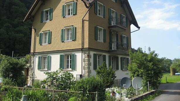 Una casa de varios pisos con un tejado a dos aguas pronunciado, numerosas ventanas con contraventanas y una base de piedra.