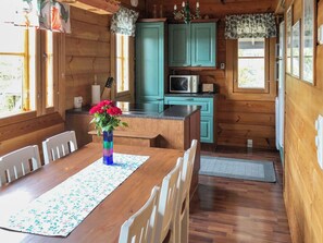 Interior de una cabaña de madera con una mesa de comedor, sillas, un florero, armarios color turquesa, un microondas y una ventana.