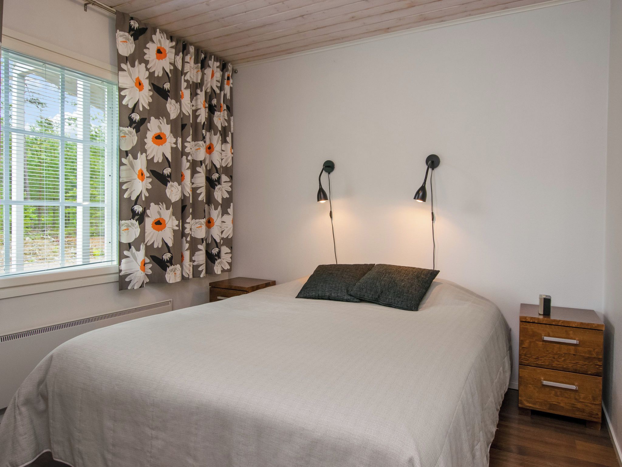 Un dormitorio con una cama, dos mesitas de noche, una ventana con persianas y una lámpara de pared.
