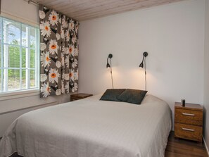 A bedroom with a bed, two bedside tables, a window with blinds, and a wall-mounted lamp.