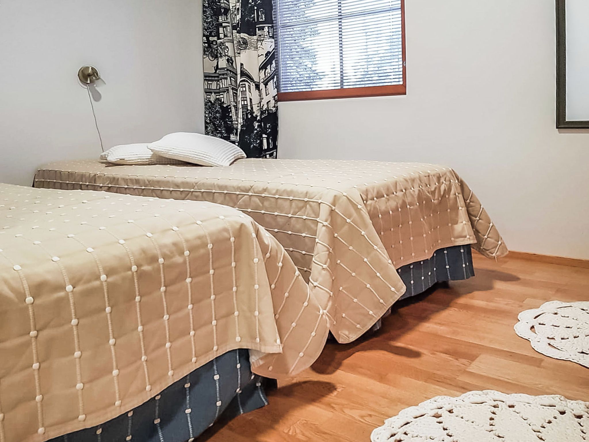 A room with two beds, each with a beige bedspread and dark blue skirted legs. There is a window with blinds, a wall-mounted lamp, and a tapestry with a cityscape design.