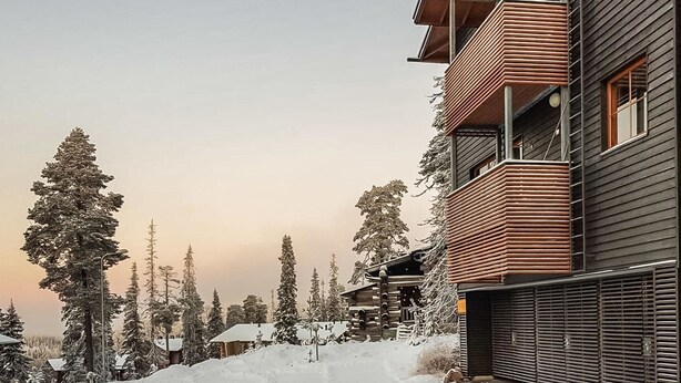 Un edificio moderno de varios pisos con fachada de madera, rodeado de árboles cubiertos de nieve y un cielo despejado.