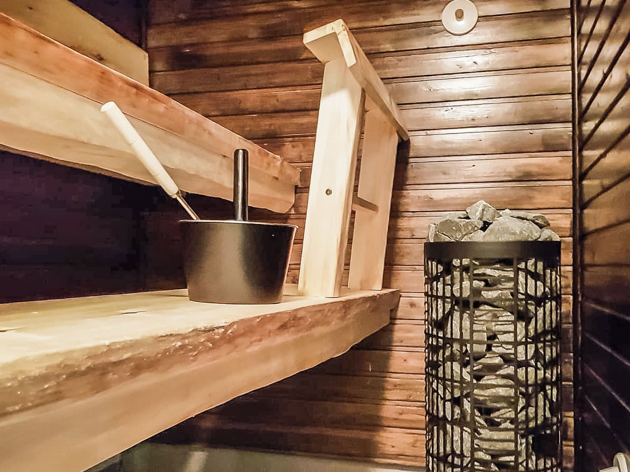 Puinen sauna, jossa on kivipenkki, metallinen ämpäri ja puulankuista rakennettu seinä.