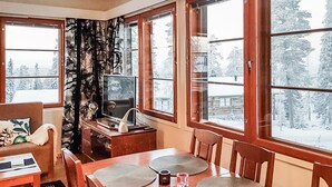 Una habitación acogedora con una mesa de comedor de madera, sillas, un televisor y grandes ventanales con vistas a un bosque nevado.
