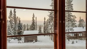 Vista a través de una ventana de madera que muestra un paisaje nevado con altos árboles de hoja perenne y una cabaña de madera.