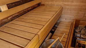 Una sala de sauna con bancos de madera, un recipiente metálico y un estufa de leña.