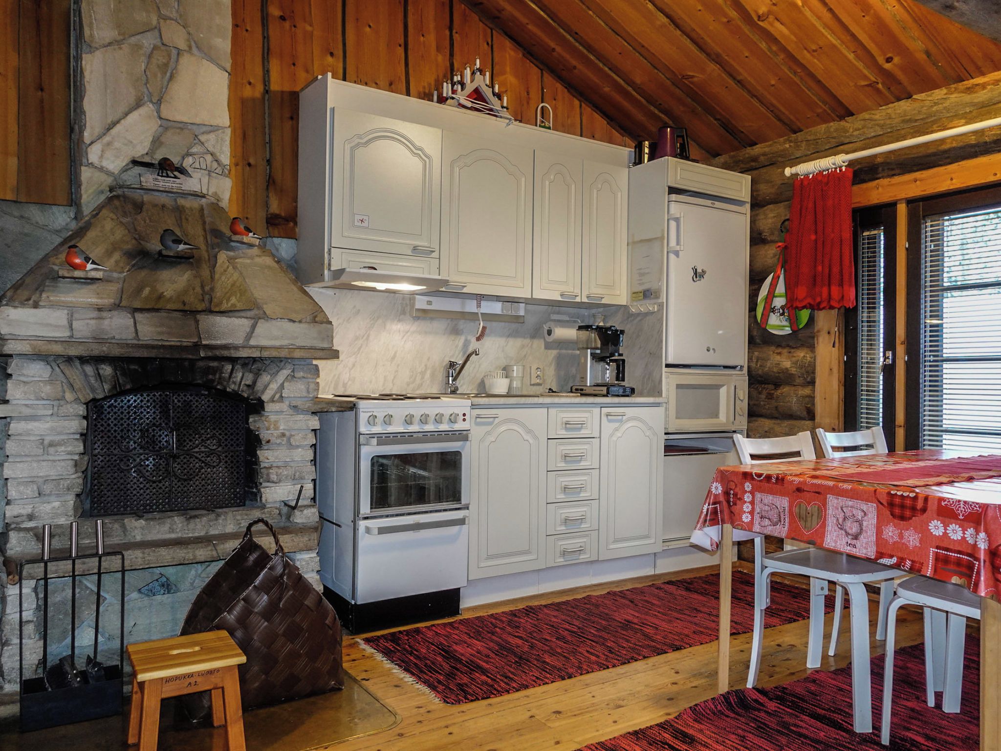 A rustic kitchen with a stone fireplace, white cabinetry, and a dining area with a red tablecloth.