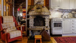 Un interior de cabaña acogedor, con una chimenea de piedra, un sillón rojo, un taburete de madera y una cocina con armarios blancos y una estufa.