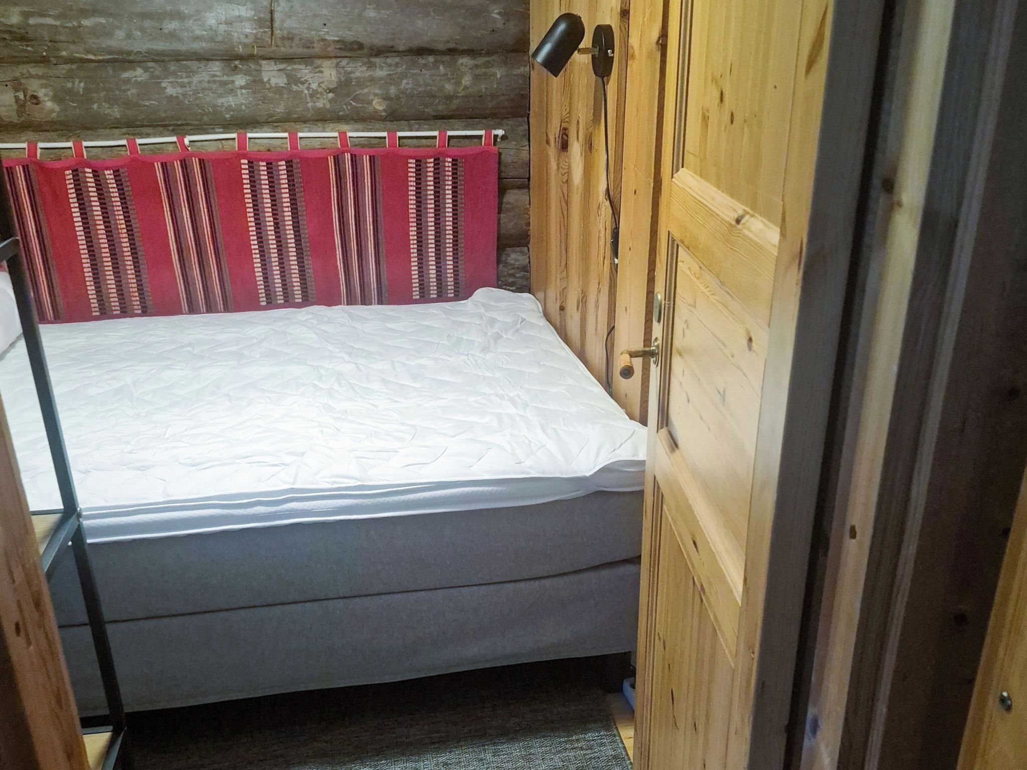 Ein kleiner, abgeschlossener Raum mit einem Bett, einer Holztür und einem Fenster mit roten Vorhängen.