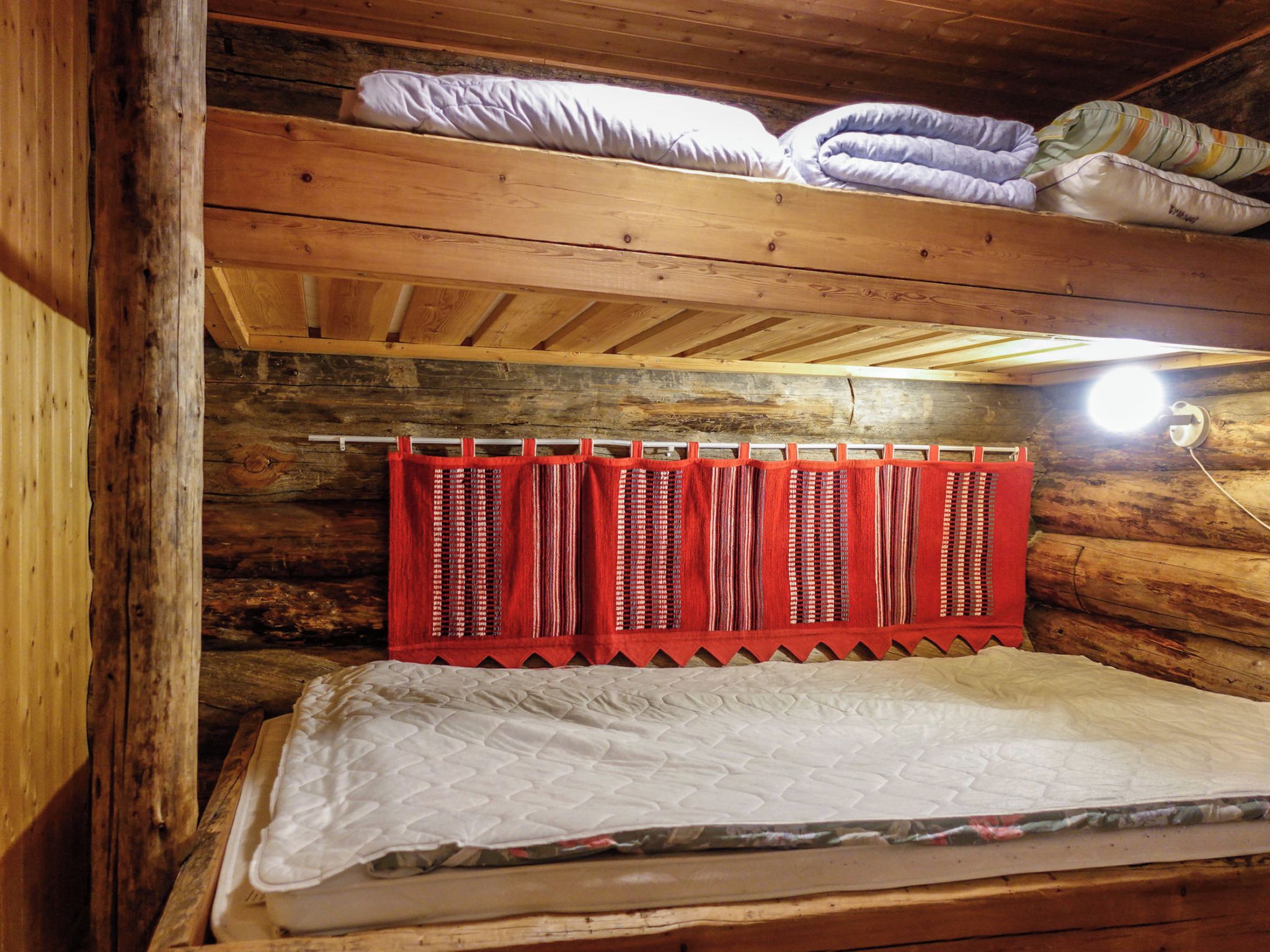 A bunk bed with a mattress and pillows, a wooden ceiling, and a red curtain.