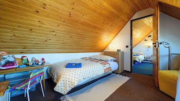 A bedroom with a sloped ceiling, a bed with patterned bedding, a desk with toys, a chair, and a door leading to another room.